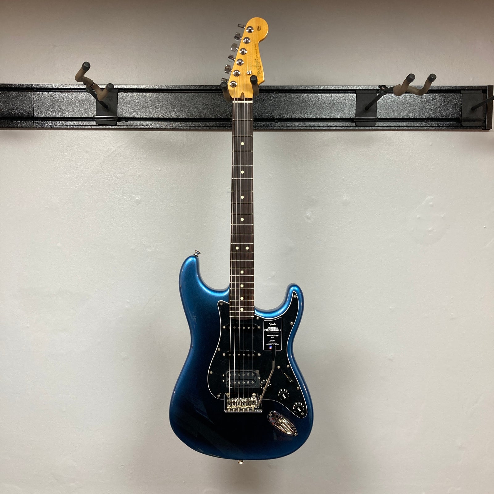 A Fender American Professional II Stratocaster HSS in Dark Night with case, featuring a black pickguard, photographed against a plain light gray wall. A Fender American Professional II Stratocaster HSS in Dark Night with case, featuring a black pickguard, photographed against a plain light gray wall.