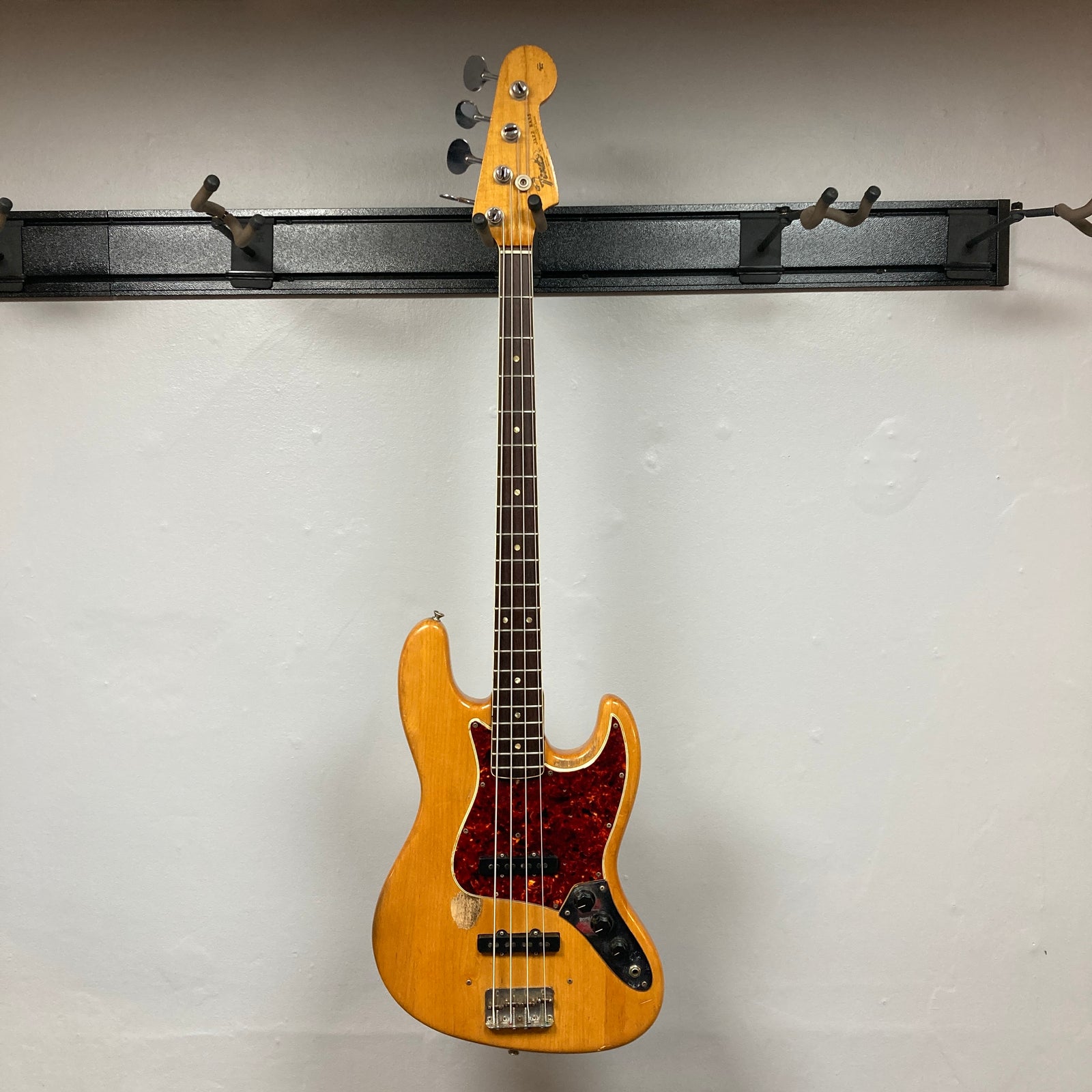 A 1966 Fender Jazz Bass Natural Refinish by Fender, featuring a red tortoiseshell pickguard, black control knobs, and original pickups, hangs against a plain light gray wall. A 1966 Fender Jazz Bass Natural Refinish by Fender, featuring a red tortoiseshell pickguard, black control knobs, and original pickups, hangs against a plain light gray wall.