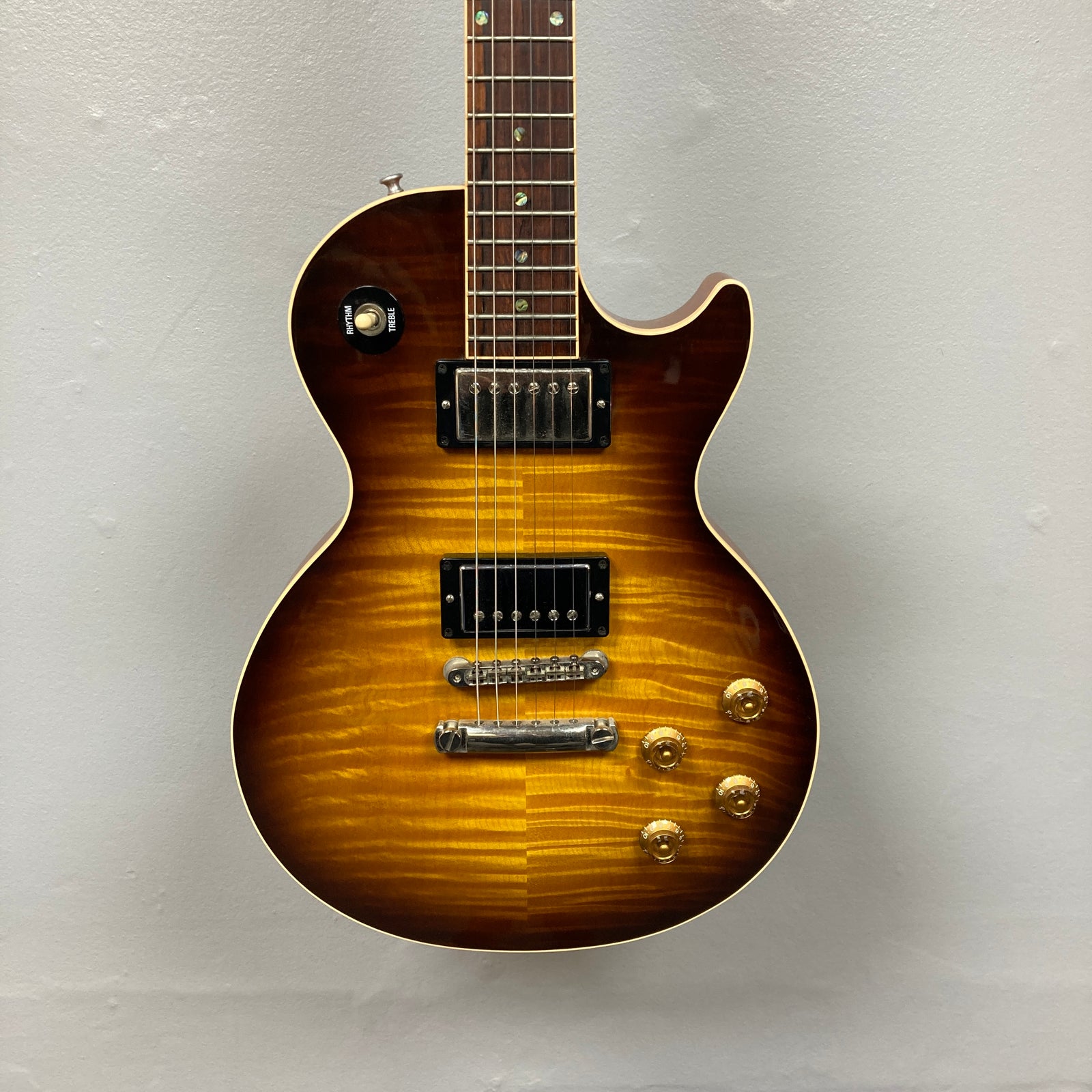 A Gibson Custom Shop Class 5 Les Paul electric guitar with a flame maple top, two humbuckers, four gold control knobs, and a three-way toggle switch hangs against a plain light gray wall. A Gibson Custom Shop Class 5 Les Paul electric guitar with a flame maple top, two humbuckers, four gold control knobs, and a three-way toggle switch hangs against a plain light gray wall.