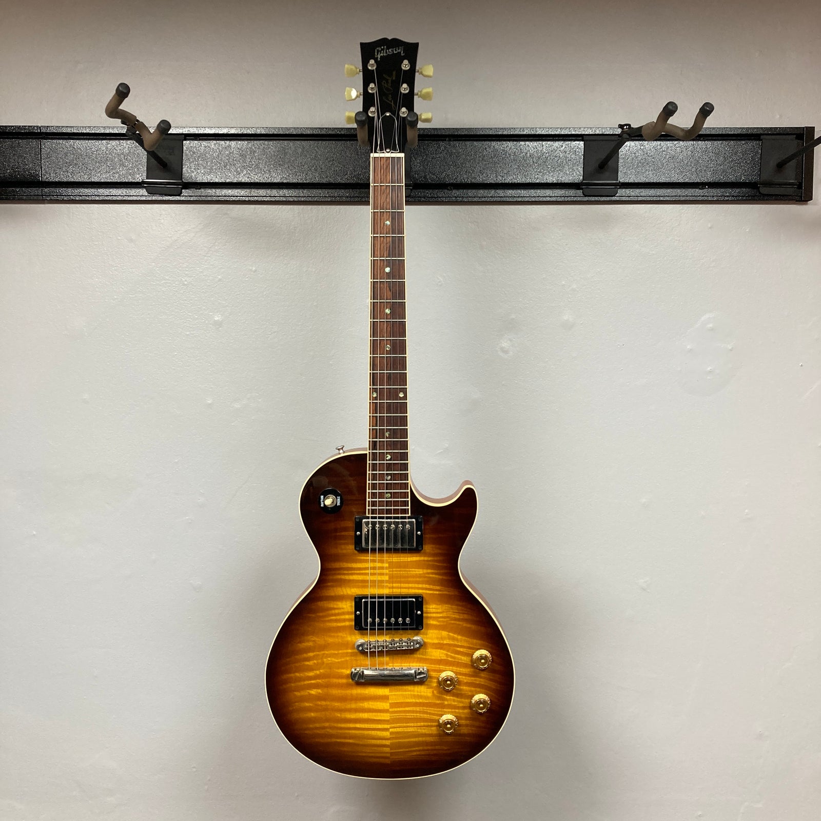 A Gibson Custom Shop Class 5 Les Paul electric guitar with a flame maple top, two humbuckers, four gold control knobs, and a three-way toggle switch hangs against a plain light gray wall. A Gibson Custom Shop Class 5 Les Paul electric guitar with a flame maple top, two humbuckers, four gold control knobs, and a three-way toggle switch hangs against a plain light gray wall.