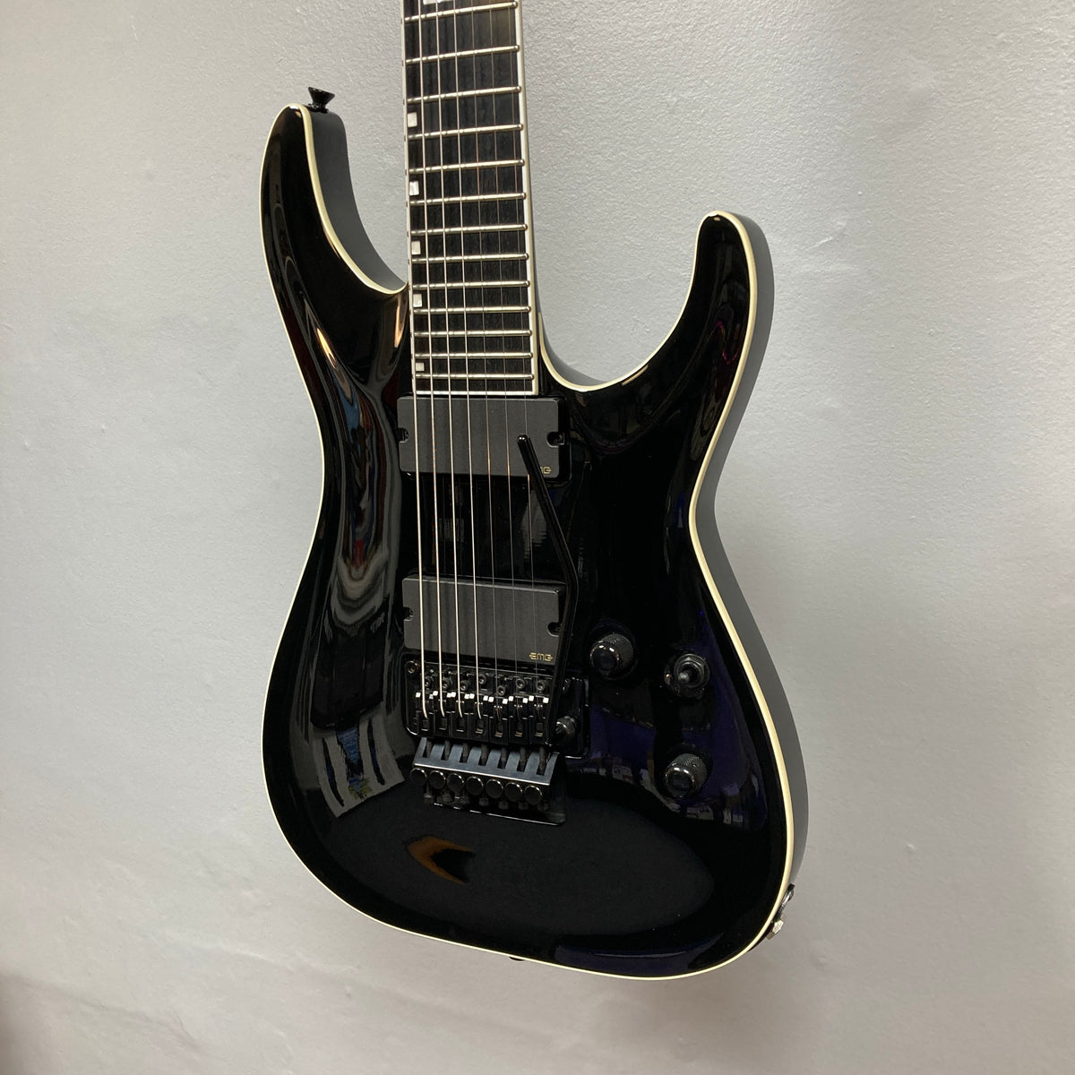 An ESP E-II Horizon FR-7 Black 7-string electric guitar with a gloss finish, white binding, humbuckers, Floyd Rose bridge, and three knobs hangs against a white wall. Comes with case.