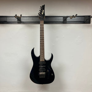 An Ibanez Prestige RG1570 Cosmic Blue electric guitar with case sits on a wall-mounted rack against a plain white wall, highlighting its vivid blue finish and signature Wizard Prestige neck.