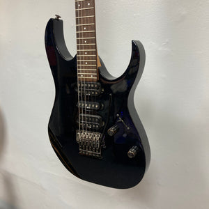 An Ibanez Prestige RG1570 Cosmic Blue electric guitar with a double-cutaway body and tremolo bridge hangs on a white wall.