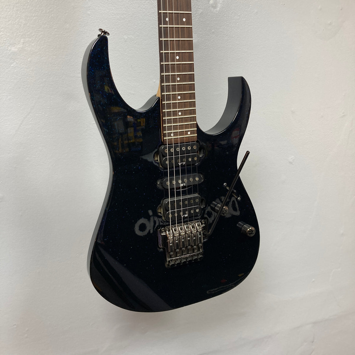 An Ibanez Prestige RG1570 electric guitar in Cosmic Blue with a case, featuring a double-cutaway body, three pickups, and a Wizard Prestige neck, hangs on a white wall.