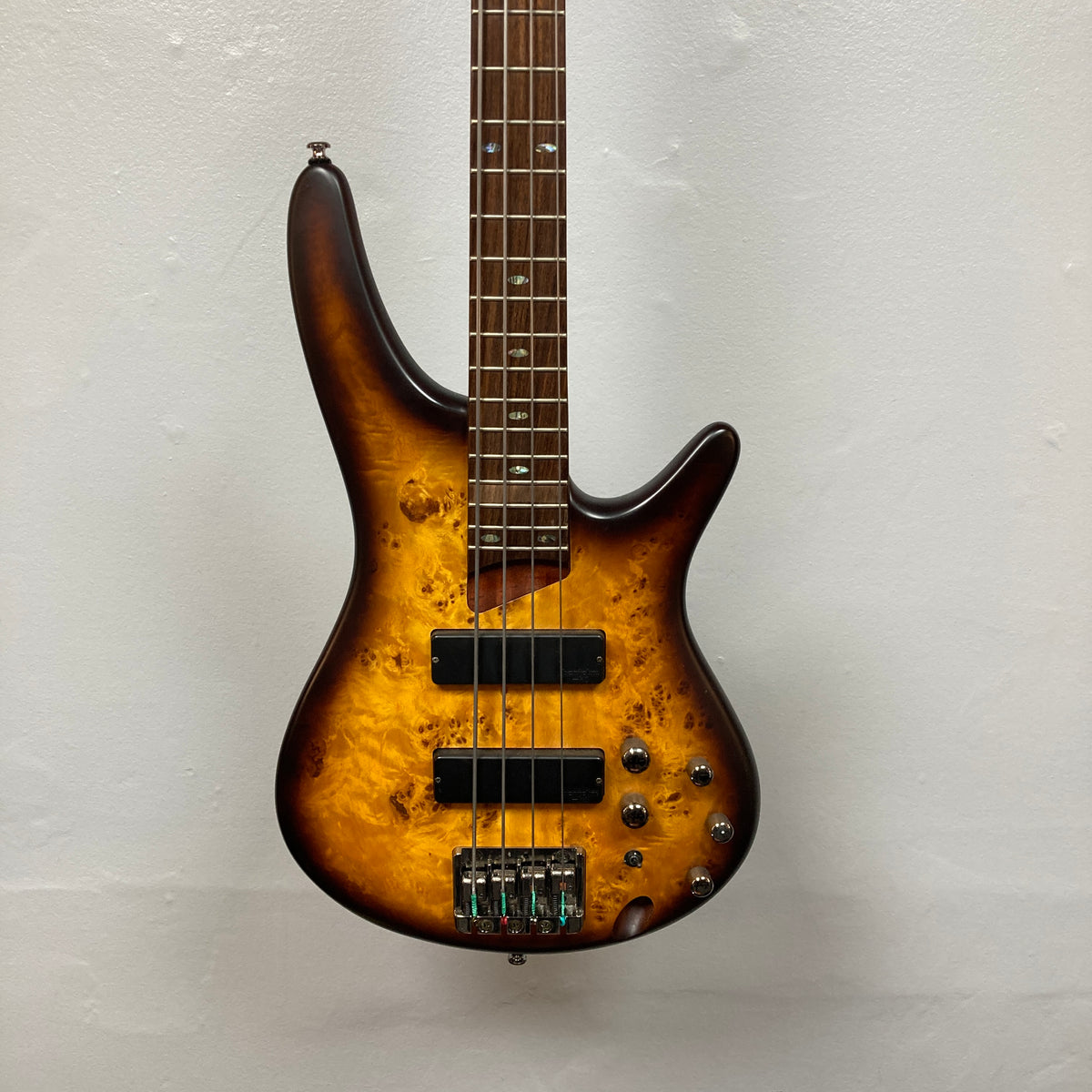An Ibanez SDGR SR500PB 4-string bass guitar with a sunburst finish, flatwound strings, dual pickups, and multiple control knobs hangs on a plain white wall. Includes hard case.
