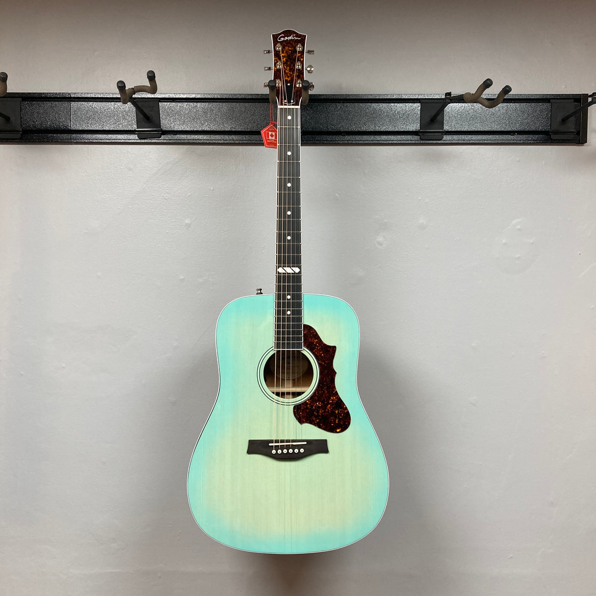 A Godin Imperial Laguna Blue GT EQ SF Acoustic Guitar w/Gigbag hangs on a wall-mounted holder against a plain white wall.