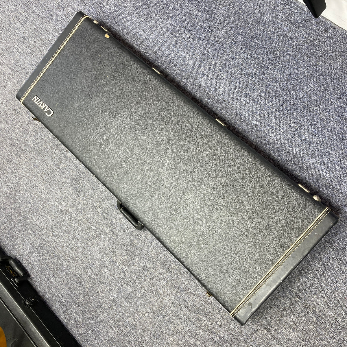 A closed black Carvin guitar case with white stitching, likely holding a 1992 Carvin Ultra V USA – Kahler Steeler Trem, MJ-Wound Seymour Duncan DCJ electric guitar, rests on a gray carpeted floor.