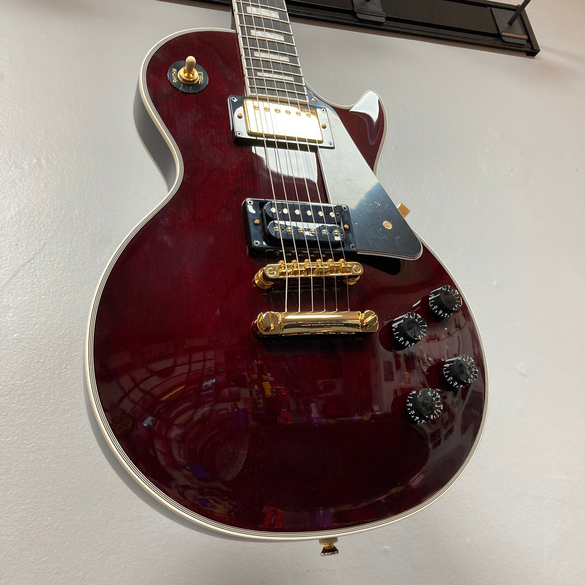 A close-up of the Epiphone Jerry Cantrell Wino Les Paul Custom in Wine Red with gold hardware and black accents, hanging on a wall. Includes a custom hardshell case for safe storage and transport.