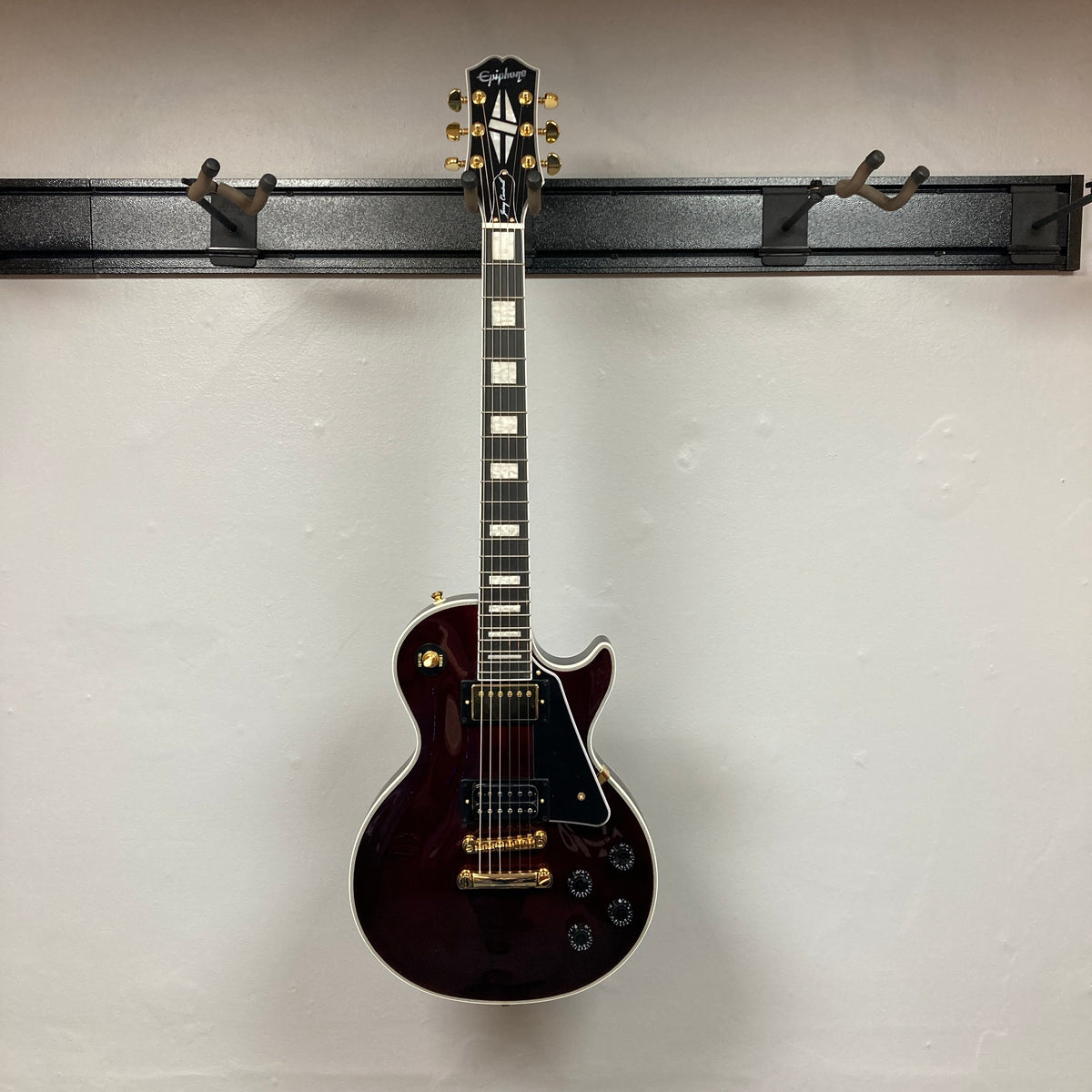 An Epiphone Jerry Cantrell Wino Les Paul Custom electric guitar in wine red with gold hardware and white trim hangs on a wall-mounted rack against a plain white background. Comes with case.