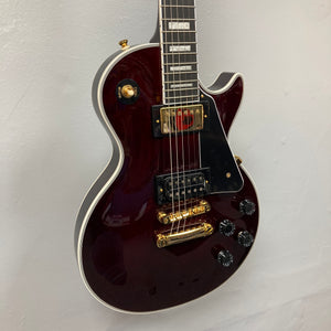 An Epiphone Jerry Cantrell Wino Les Paul Custom in Wine Red with gold hardware, black pickguard, and white trim hangs on a plain white wall.