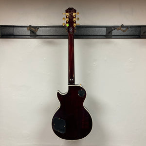 An Epiphone Jerry Cantrell Wino Les Paul Custom in Wine Red with gold tuning pegs and white trim, shown from the back, hangs on a wall-mounted rack. Case included.