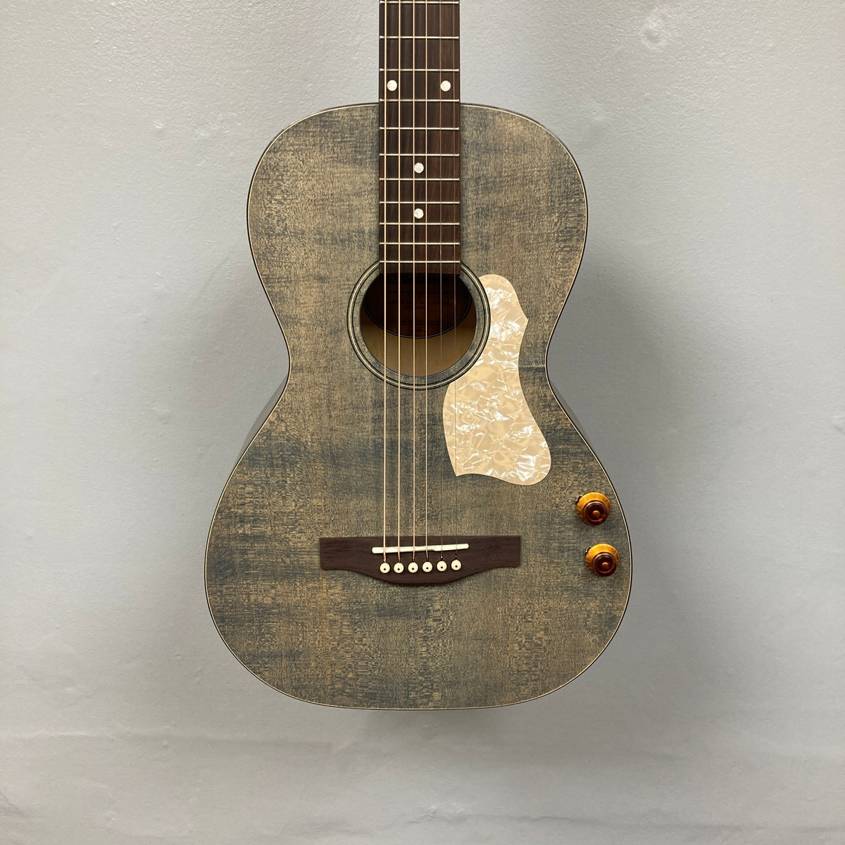 The Art & Lutherie Roadhouse Denim Blue Q-Discrete, a parlor acoustic guitar with a white pickguard, two control knobs, and six strings, hangs against a light gray wall.