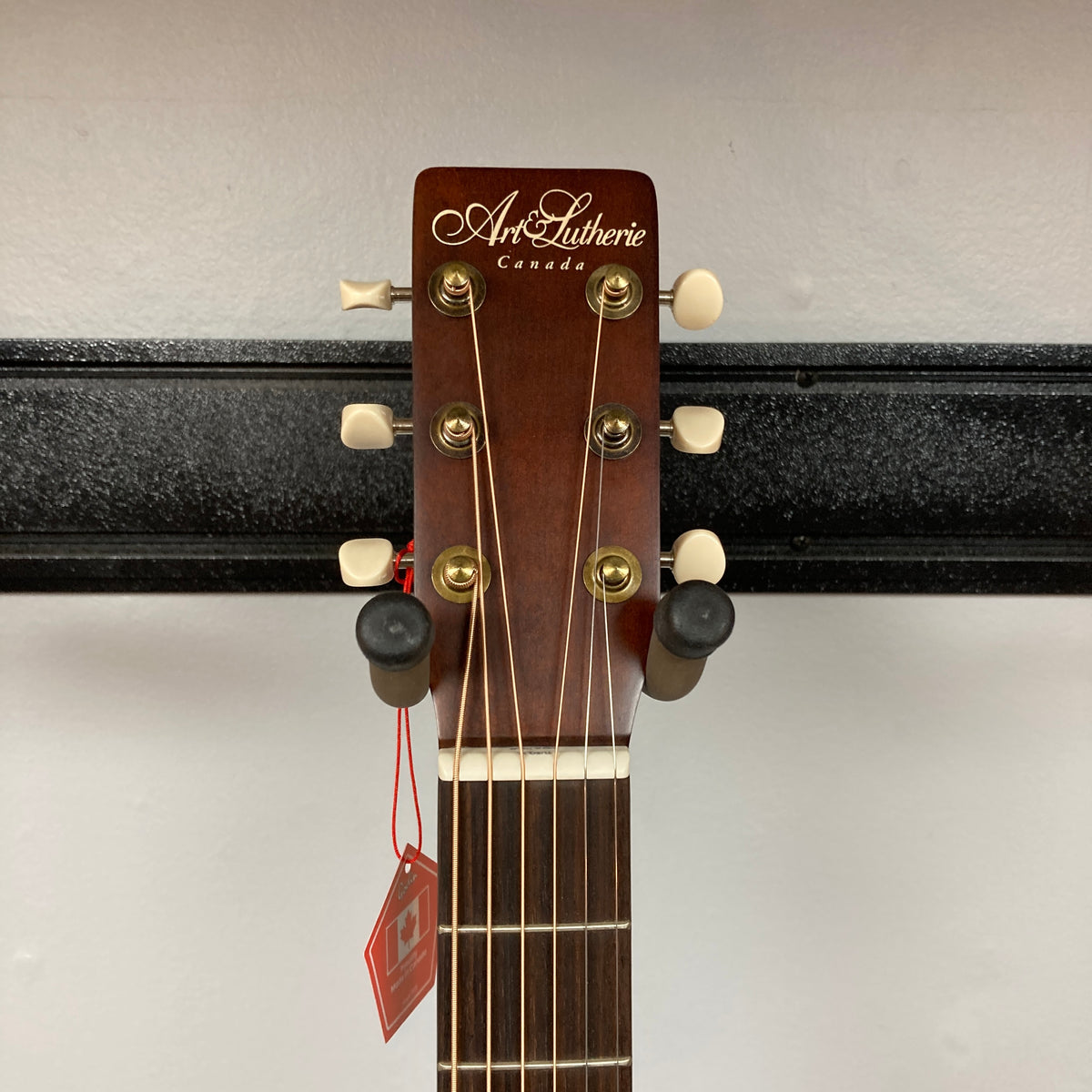 The headstock of the Art & Lutherie Roadhouse Denim Blue Q-Discrete acoustic guitar features tuning pegs, strings, and a red tag, shown against a white wall and black rack.