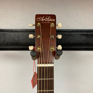 The headstock of the Art & Lutherie Roadhouse Denim Blue Q-Discrete acoustic guitar features tuning pegs, strings, and a red tag, shown against a white wall and black rack.