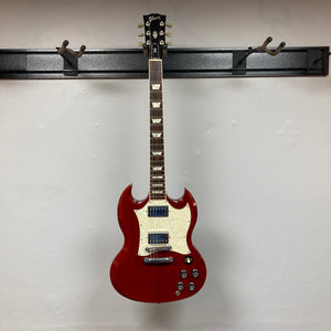 A Gibson SG Standard Heritage Cherry yr 2000 Consign electric guitar by Gibson, featuring a white pickguard, hangs on a wall-mounted rack against a plain white wall, highlighting its USA-made craftsmanship.