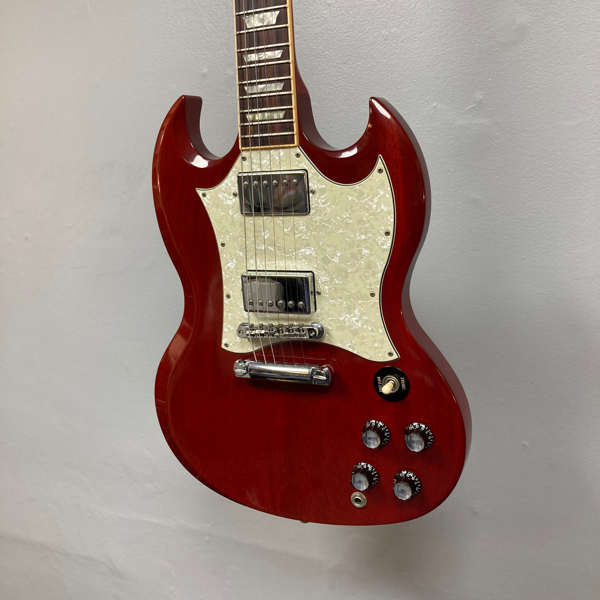 A Gibson SG Standard Heritage Cherry yr 2000 Consign electric guitar with a white pickguard, four control knobs, and two humbucker pickups hangs against a plain white wall.