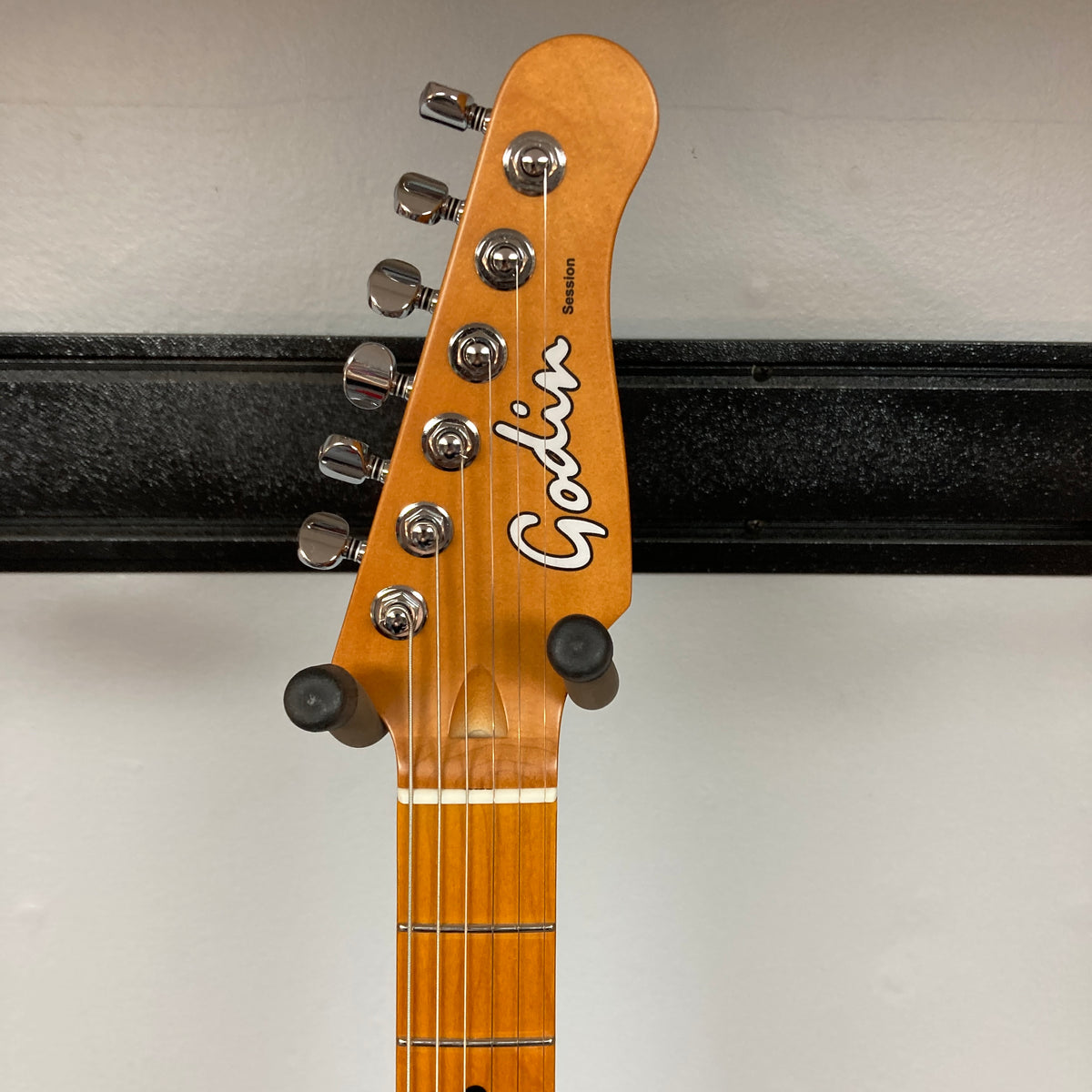 Close-up of a Godin Session T-Pro Arctik Blue MN SF with Bag headstock, showing six tuning pegs and the Godin logo on a wood finish, plus part of a wall-mounted holder. Features HSS Seymour Duncan pickups.