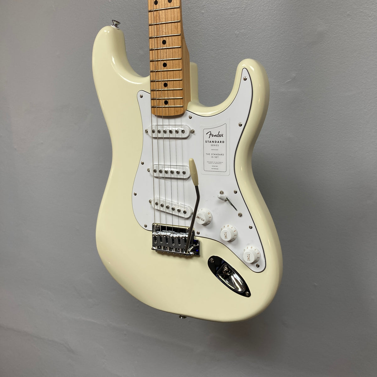 A Fender Standard Stratocaster Olympic White Refurb, featuring a maple neck and white pickguard, hangs against a plain gray wall.