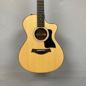 A used Taylor 212ce Plus w/Gigbag acoustic guitar by Taylor, with a natural finish and black pickguard, is shown against a plain light gray wall.
