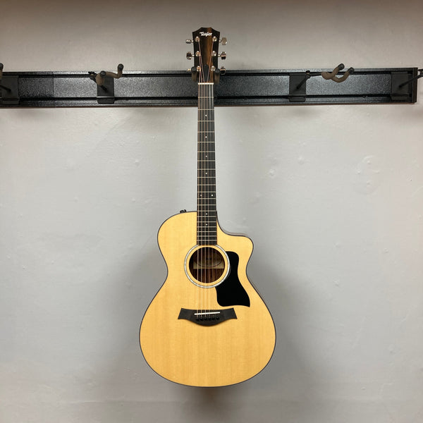 A used Taylor 212ce Plus w/Gigbag acoustic guitar by Taylor, with a natural finish and black pickguard, is shown against a plain light gray wall.
