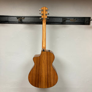 A used Taylor 212ce Plus w/Gigbag acoustic guitar by Taylor is displayed from the back, highlighting its natural wood grain finish on a wall hanger against a plain light-colored wall.