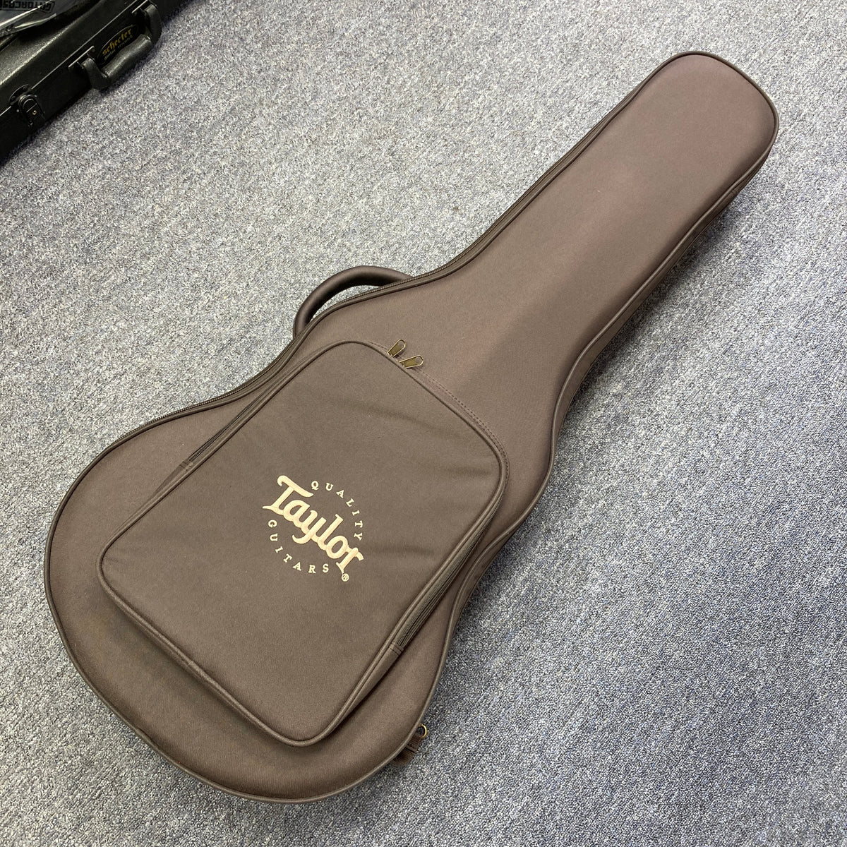 A Taylor 212ce Plus acoustic guitar gig bag with a front pocket, in brown, is on a gray carpeted floor.