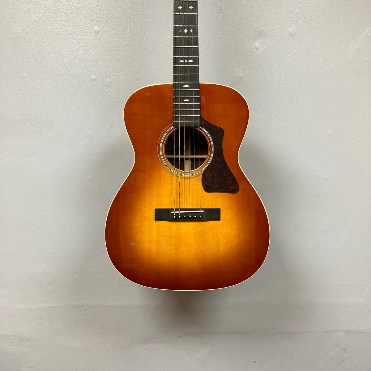 The Guild Gad-30reasb w/Case, an orchestra-size acoustic-electric guitar with a sunburst finish, is hanging on a plain, light-colored wall and features a Fishman Matrix pickup for exceptional amplified sound.