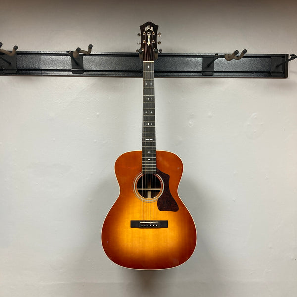The Guild Gad-30reasb w/Case, an orchestra-size acoustic-electric guitar with a sunburst finish, is hanging on a plain, light-colored wall and features a Fishman Matrix pickup for exceptional amplified sound.