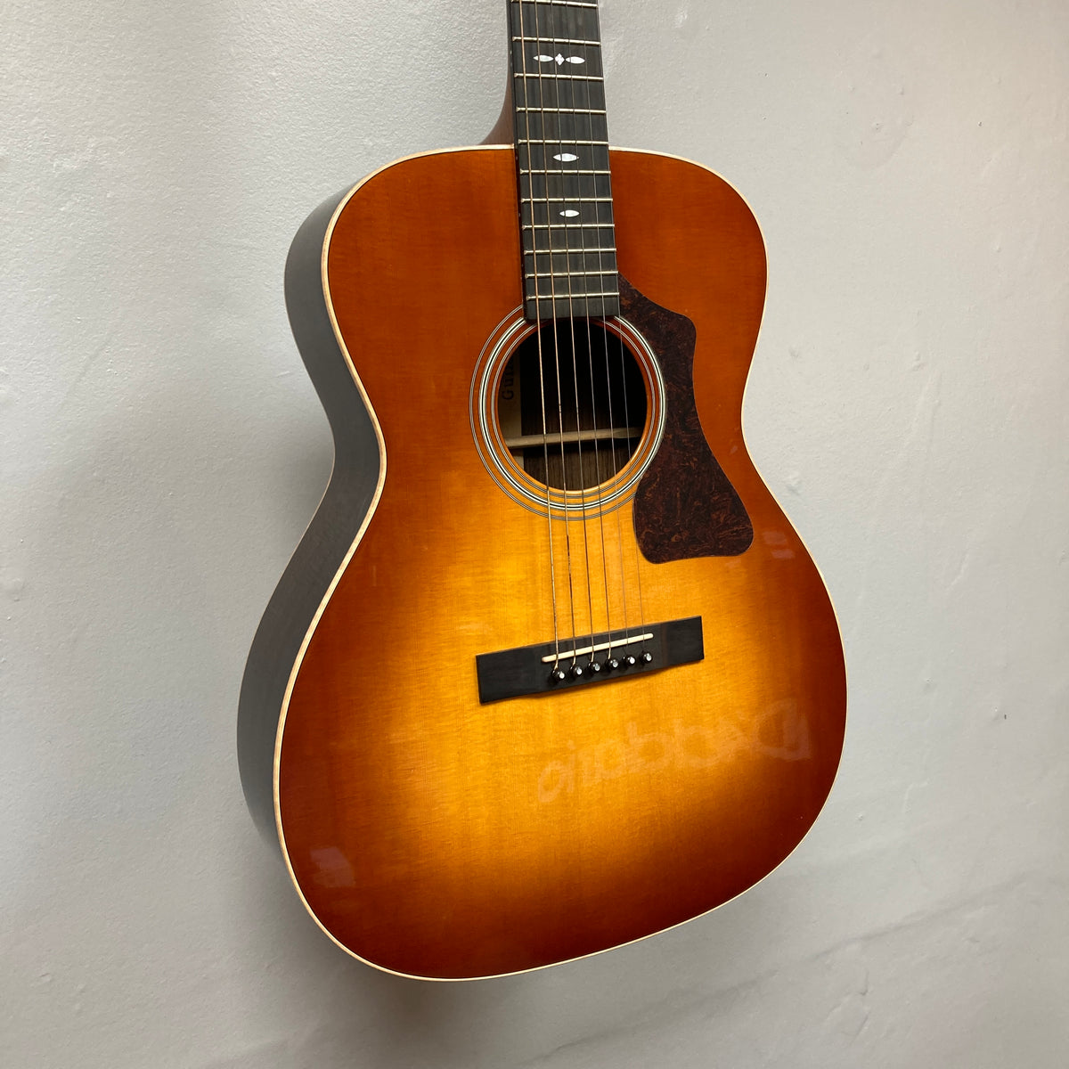 A Guild Gad-30reasb acoustic guitar with a sunburst finish, crafted from solid Indian rosewood, is hanging on a white wall, displaying its front body and fretboard.