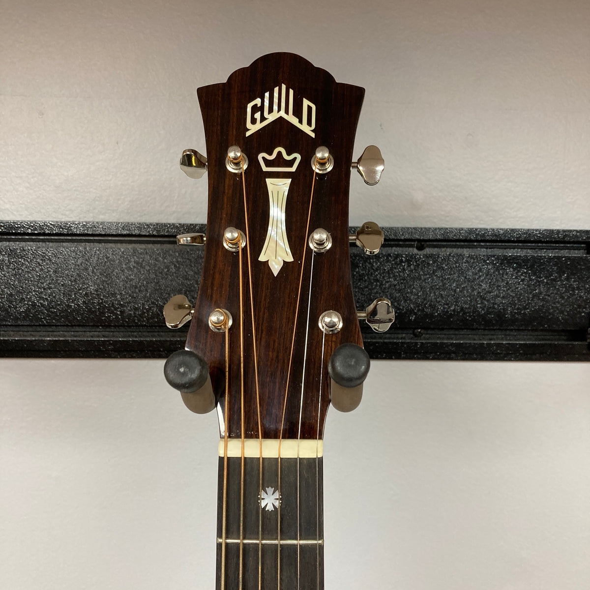 Headstock of a Guild Gad-30reasb acoustic-electric guitar, crafted from solid Indian rosewood, featuring tuning pegs and a decorative inlay, mounted on a wall hanger.