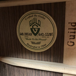 A close-up of a Guild Gad-30REASB w/Case guitar label inside the body displays the model GAD-30REASB, serial number GAD-12291, and the phrase "Made To Be Played," indicating this orchestra-sized acoustic-electric guitar by Guild was handcrafted in China with solid Indian rosewood back and sides, and is equipped with a premium Fishman Matrix pickup.