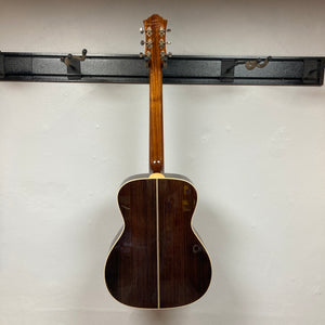 The Guild Gad-30reasb w/Case acoustic-electric guitar by Guild is displayed hanging on a wall, viewed from the back to showcase its dark solid Indian rosewood finish and metal tuning pegs.