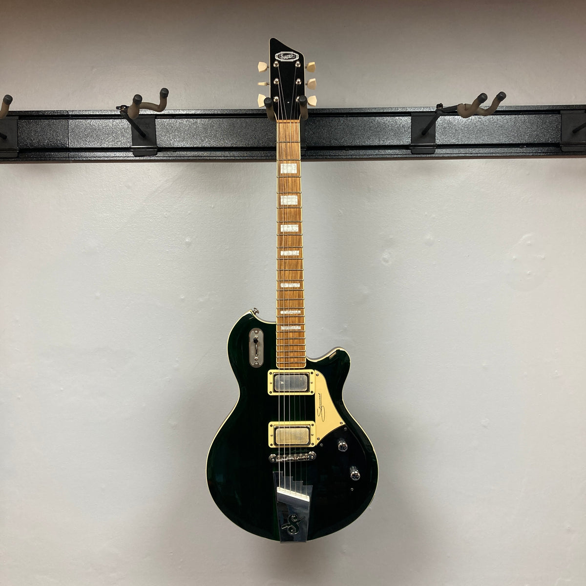 A Supro 1296 Silverwood in British Racing Green, featuring gold hardware and unique Gold Foil pickups, hangs on a wall-mounted rack. The eye-catching finish of this electric guitar stands out beautifully against the white background.