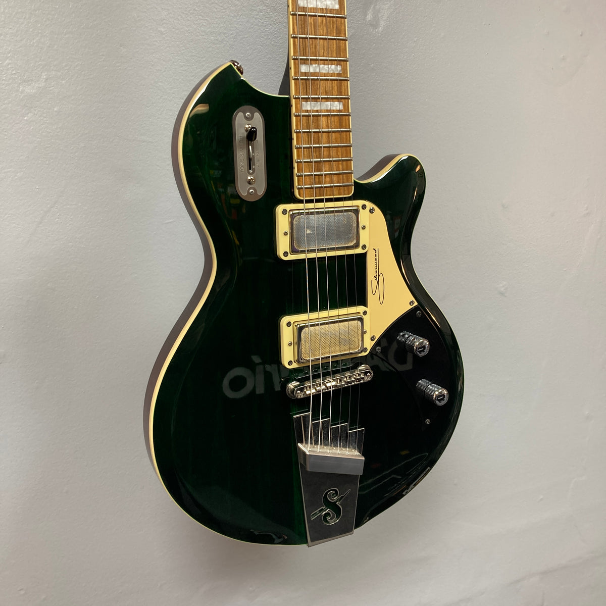 Displayed against a plain light-colored wall is the Supro 1296 Silverwood in British Racing Green, featuring cream pickups, gold hardware, and a signature on the pickguard.