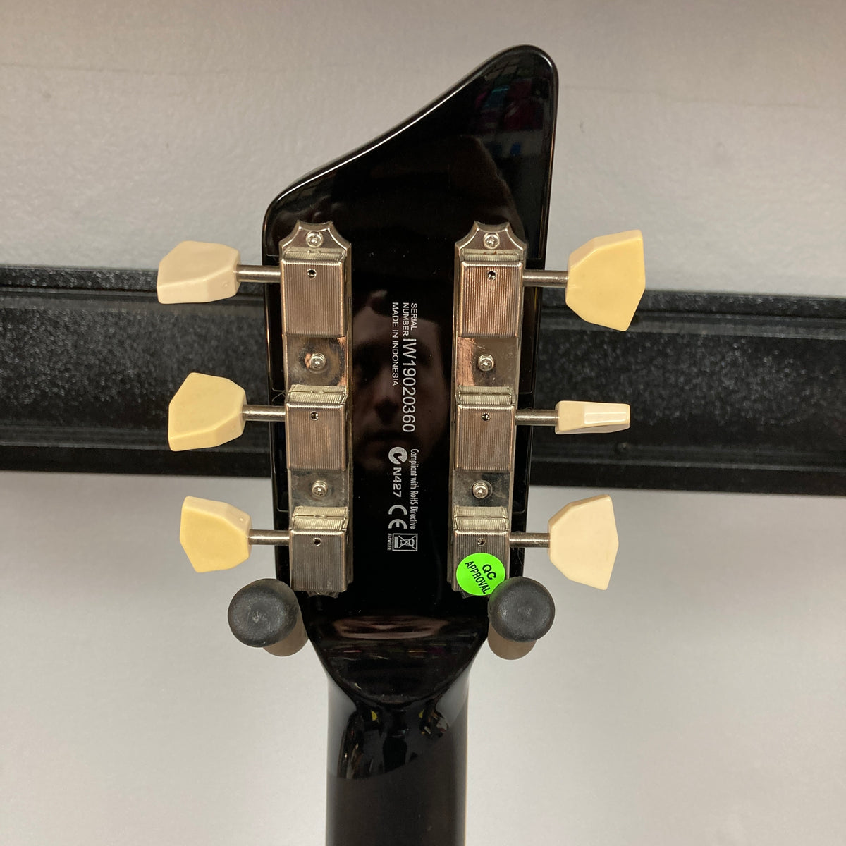 The back of a Supro 1296 Silverwood British Racing Green guitar headstock, featuring tuning pegs, a serial number, a manufacturer's label, and a green "QC OK" sticker alongside a wall mount holder—elements that highlight the meticulous craftsmanship of Supro's iconic model.