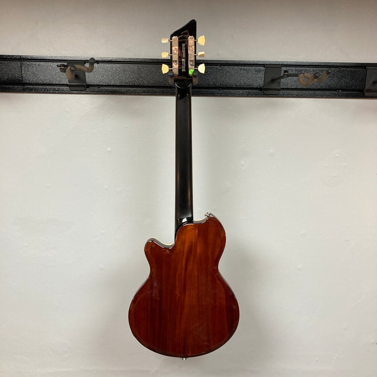 A Supro 1296 Silverwood electric guitar in British Racing Green, complete with gig bag, hangs on a black wall-mounted rack and is photographed from the back against a plain white wall, showcasing the elegant styling characteristic of Supro instruments.