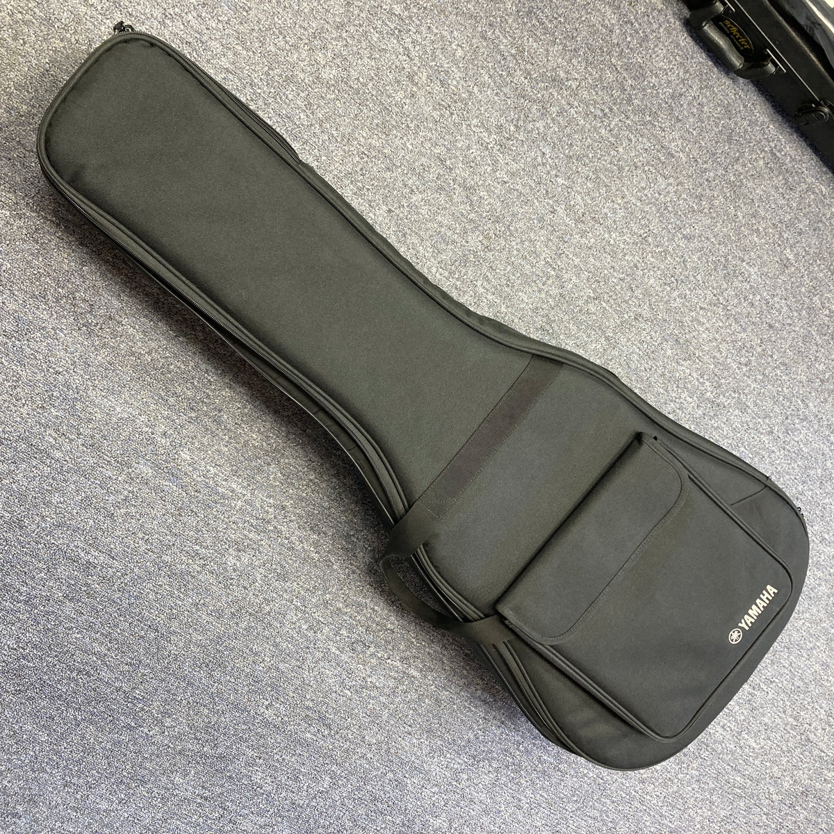 A Yamaha A3M ARE Vintage Natural Acoustic Guitar by Yamaha, featuring a solid Sitka spruce top and SRT2 pickup system, along with its included hard bag, is resting on a gray carpeted floor. The hard bag offers a convenient front pocket for storing accessories.