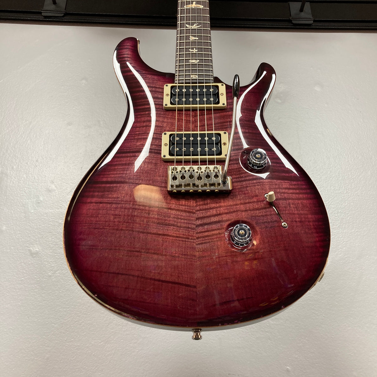 A PRS Custom 24 Violet 2011 electric guitar with a glossy finish, two 59/09 humbuckers, a tremolo bridge, and bird inlays on the fretboard is displayed against a white wall.