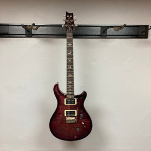 A PRS Custom 24 Violet 2011 electric guitar by PRS hangs from a black wall-mounted rack against a plain white wall.