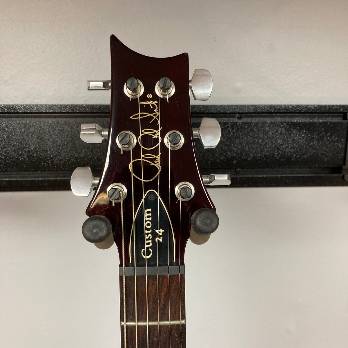 Close-up of a PRS Custom 24 Violet 2011 electric guitar headstock with signature, tuning pegs, and part of the fretboard visible; mounted on a wall hanger.