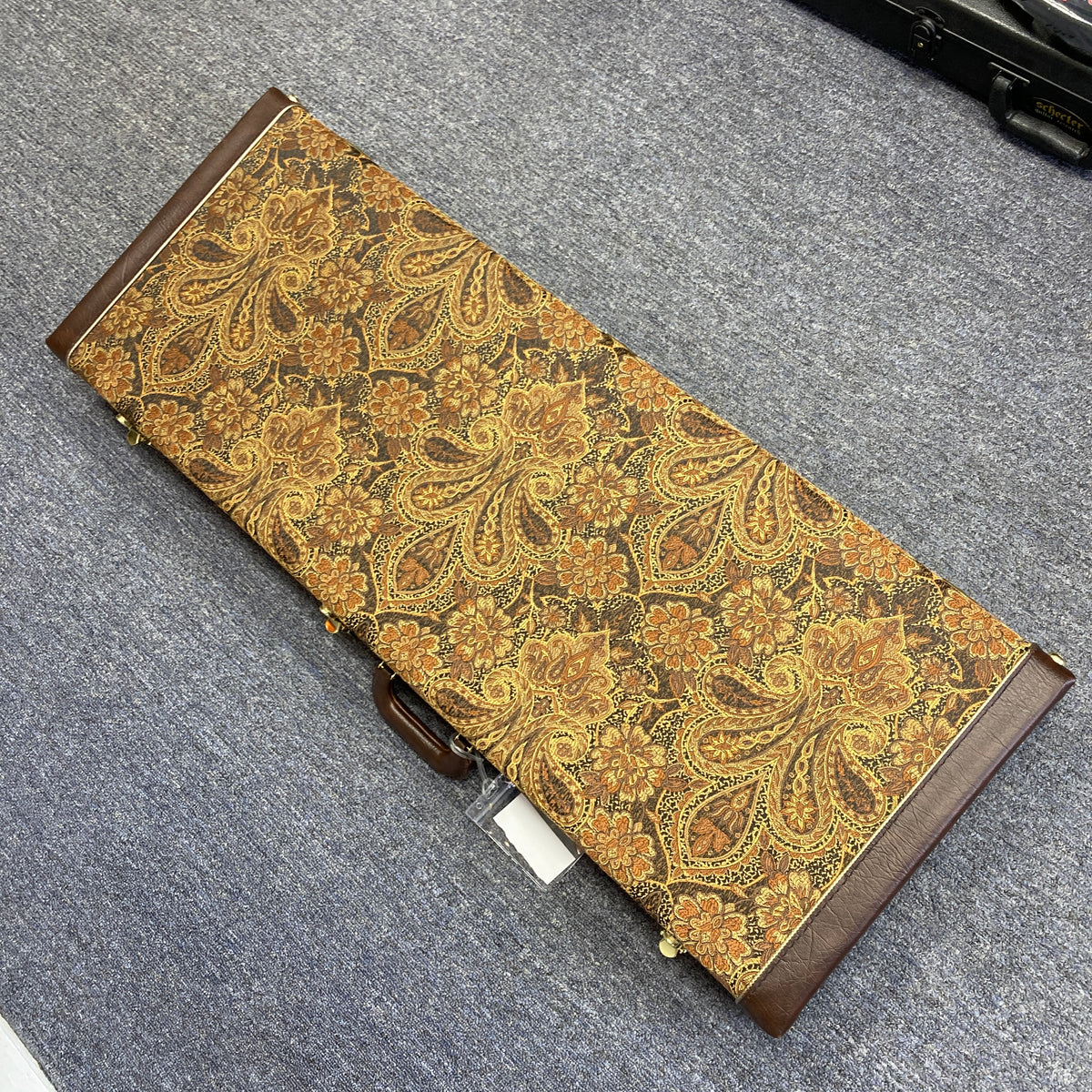 A rectangular case with a brown and gold paisley pattern rests on a gray carpeted floor. Designed to protect your PRS Custom 24 Violet 2011 electric guitar, it features brown leather accents and a sturdy handle—ideal for carrying your prized PRS instrument in style.