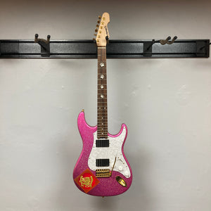 An Edwards ESP E-SN-185-TO Takayoshi Ohmura electric guitar in Twinkle Pink with a white pickguard and a red sticker is hanging on a black wall mount against a plain light-colored wall.