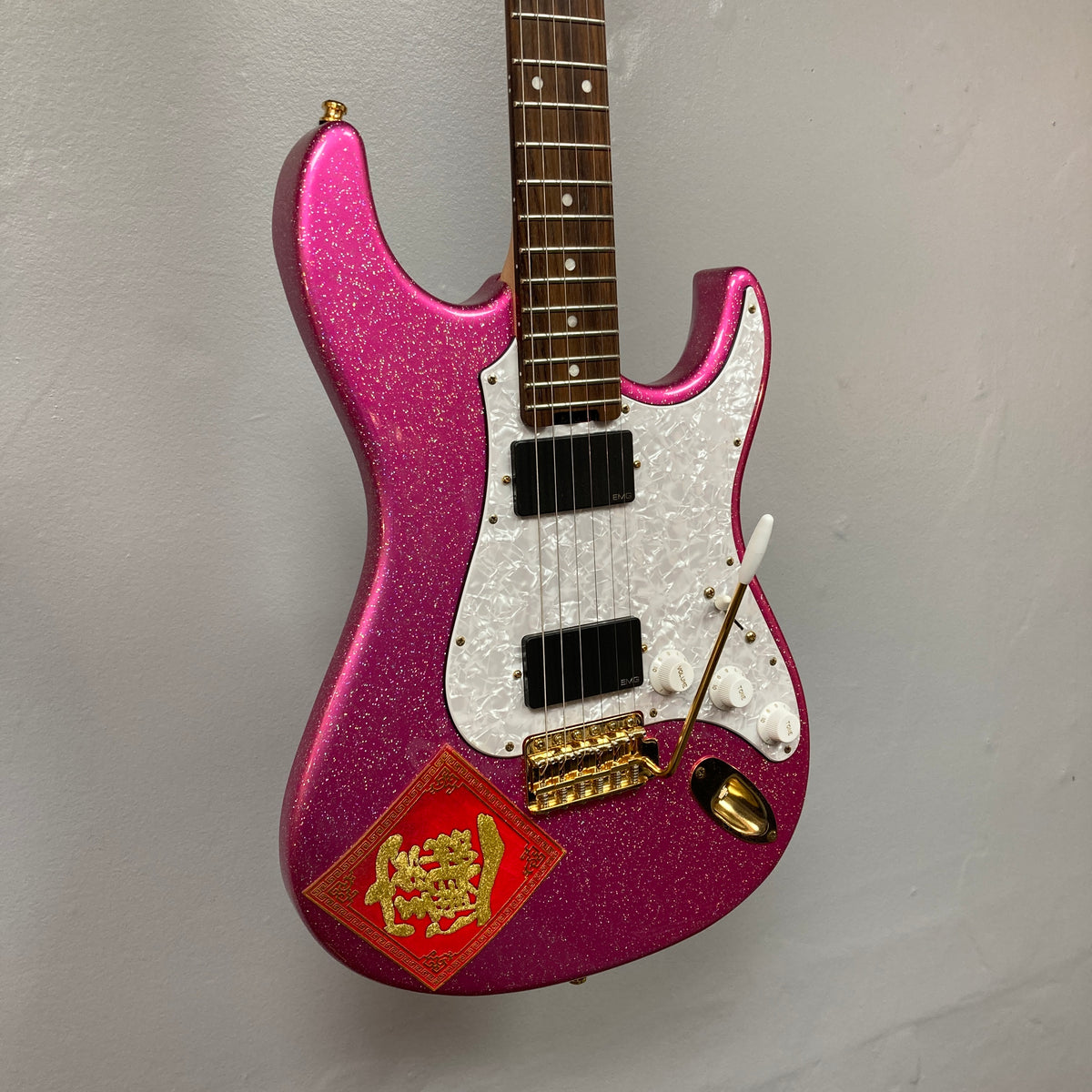 Edwards ESP E-SN-185-TO Takayoshi Ohmura Twinkle Pink electric guitar, featuring dual humbuckers, a sparkling pink glitter finish, white pickguard, gold hardware, and a red decorative sticker in the lower corner, photographed against a plain gray wall.