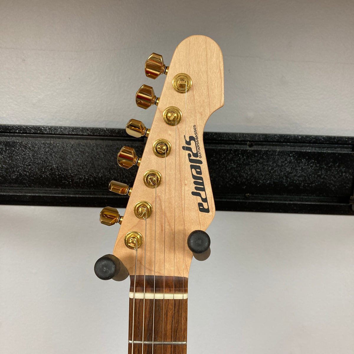 Headstock of an Edwards ESP E-SN-185-TO Takayoshi Ohmura Twinkle Pink electric guitar, featuring gold tuning pegs, mounted on a wall hanger against a plain background.