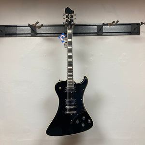 A Hagstrom Fantomen Custom Black w/case electric guitar, featuring a single cutaway, white binding, and dual Lundgren Design humbuckers, hangs on a wall-mounted guitar rack against a plain light-colored wall.