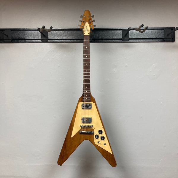 A brown 1975 Gibson Flying V electric guitar by Gibson, featuring a natural finish, distinctive V-shaped body, three control knobs, and two pickups, is displayed against a plain gray wall. Modeled after the classic vintage Flying V design, this instrument embodies the iconic style of its era and comes with a case.