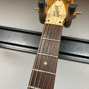 Close-up of the headstock of a 1975 Gibson Flying V w/Case by Gibson, showing part of the fretboard with six strings and a wall-mounted guitar hanger, featuring the iconic vintage Flying V design with a natural finish.