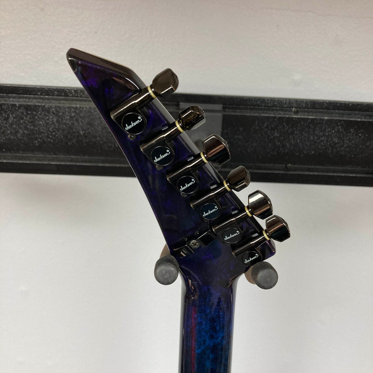 Close-up of the back of a dark electric guitar’s headstock with six Jackson-branded tuning pegs, hanging on a wall-mounted holder—this striking piece is a 1995 Jackson SL1 Soloist Custom Shop Eerie Dess Swirl w/Case.