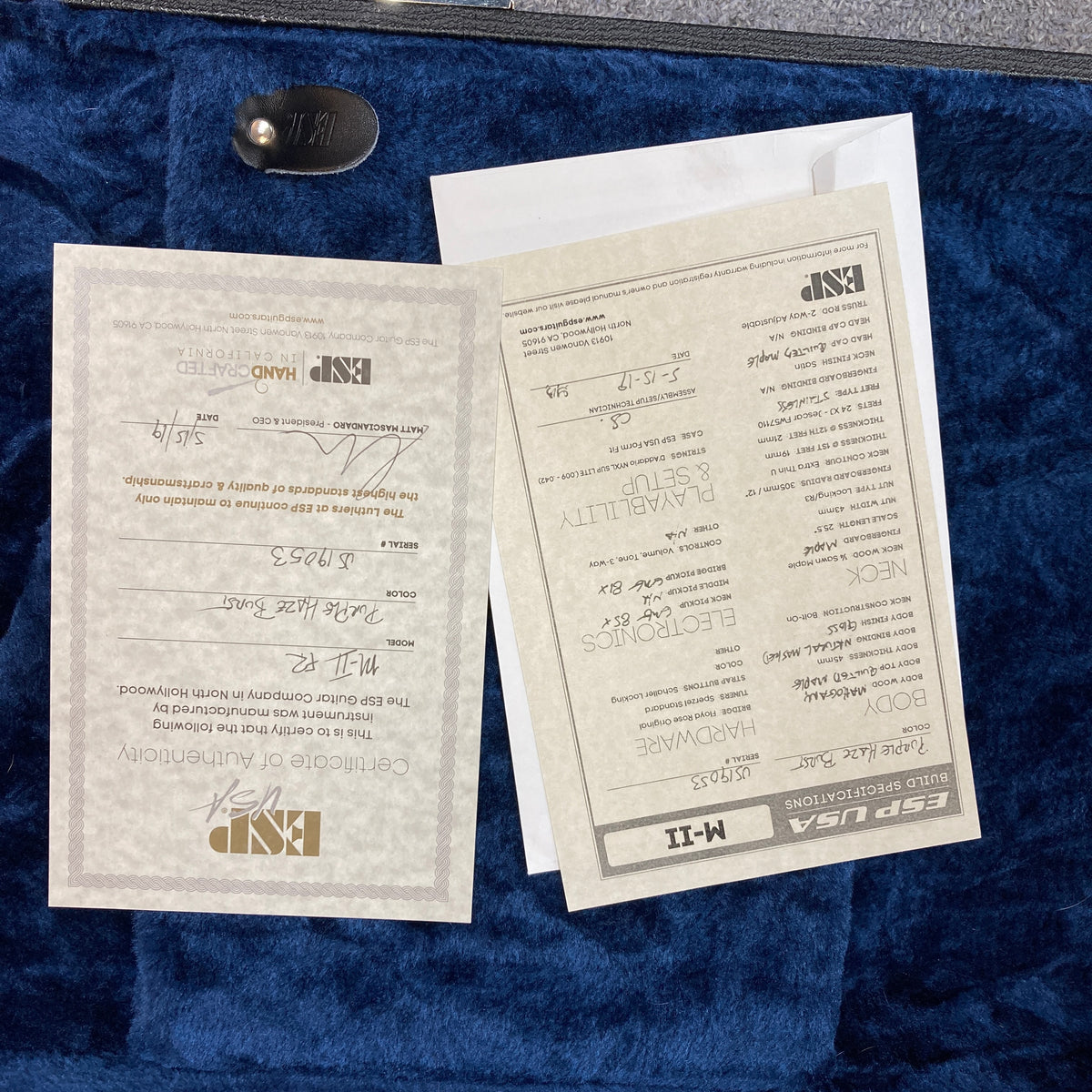 Two ESP specification and warranty cards, one of which features handwritten details, rest atop a blue velvet-lined surface—the perfect accompaniment to your American-made electric guitar, like the striking 2019 ESP USA M-II Purple Haze Burst w/Case from ESP.
