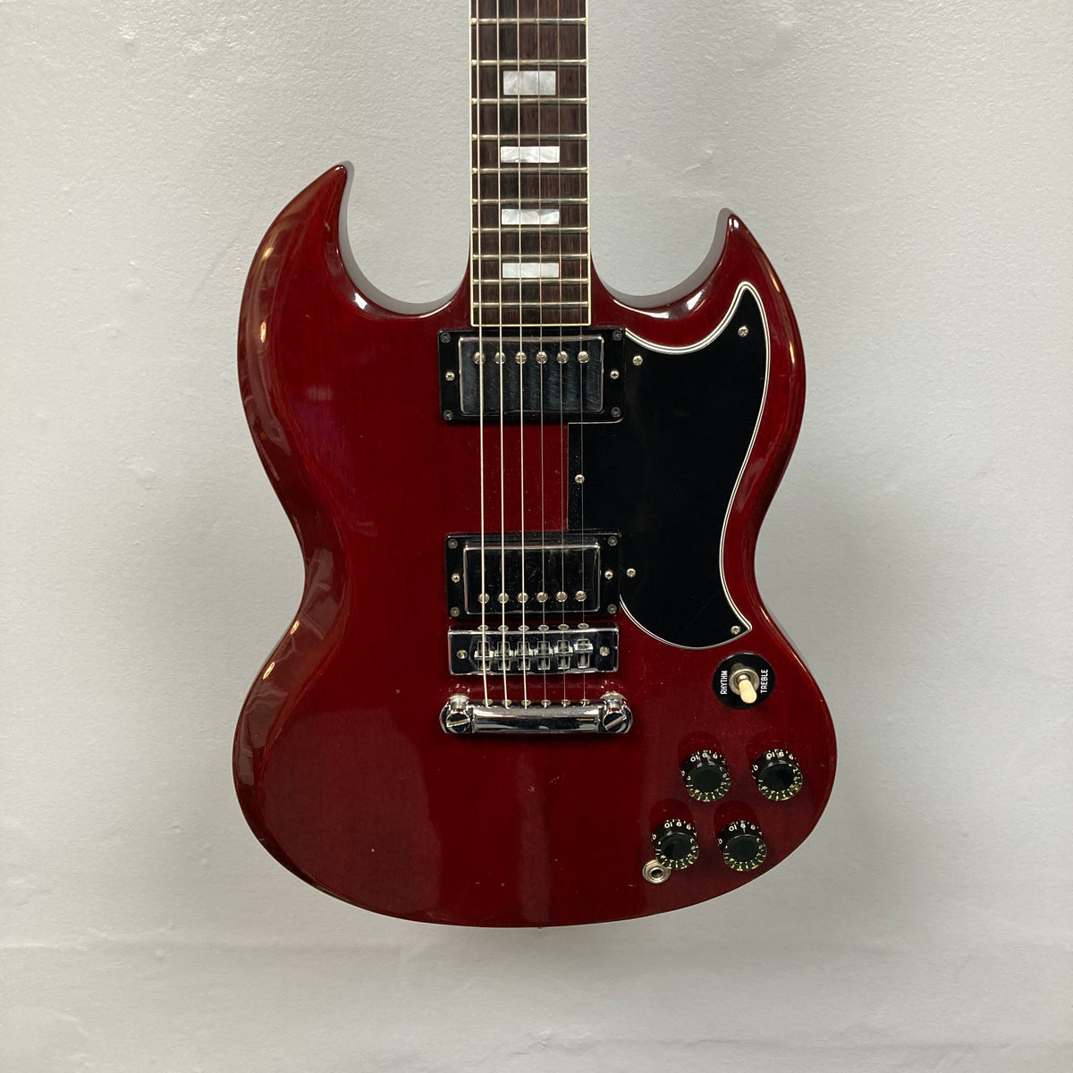 A 1977 Gibson SG Standard Cherry electric guitar by Gibson, featuring a classic double-cutaway mahogany body, two humbucker pickups, four control knobs, and a black pickguard evocative of the iconic Kalamazoo era, hangs elegantly against a plain white wall.